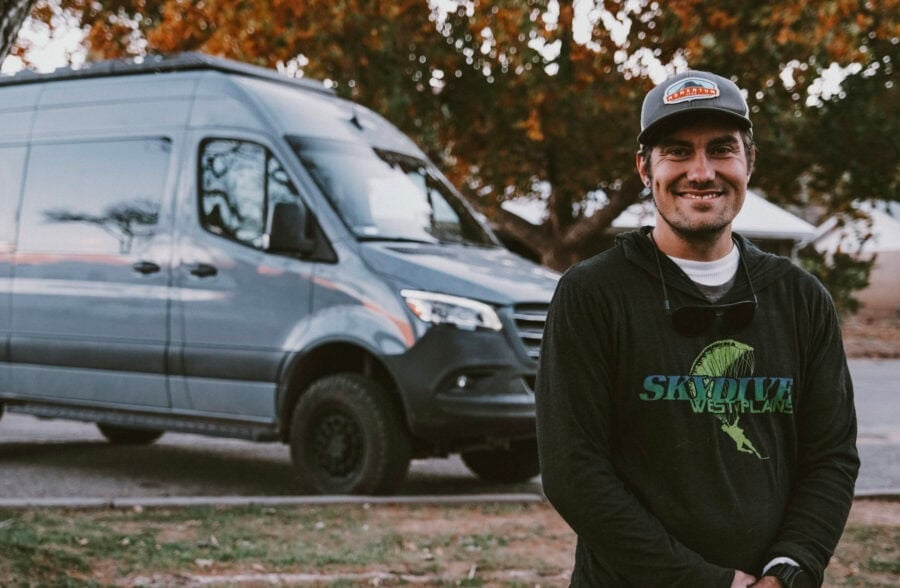 Skydiver standing next to a 4x4 Sprinter van in a scenic outdoor setting.