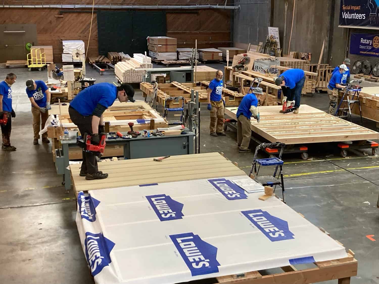 Volunteers in blue shirts framing tiny house walls and floor panels on rolling jigs inside the Hope Factory warehouse in Seattle