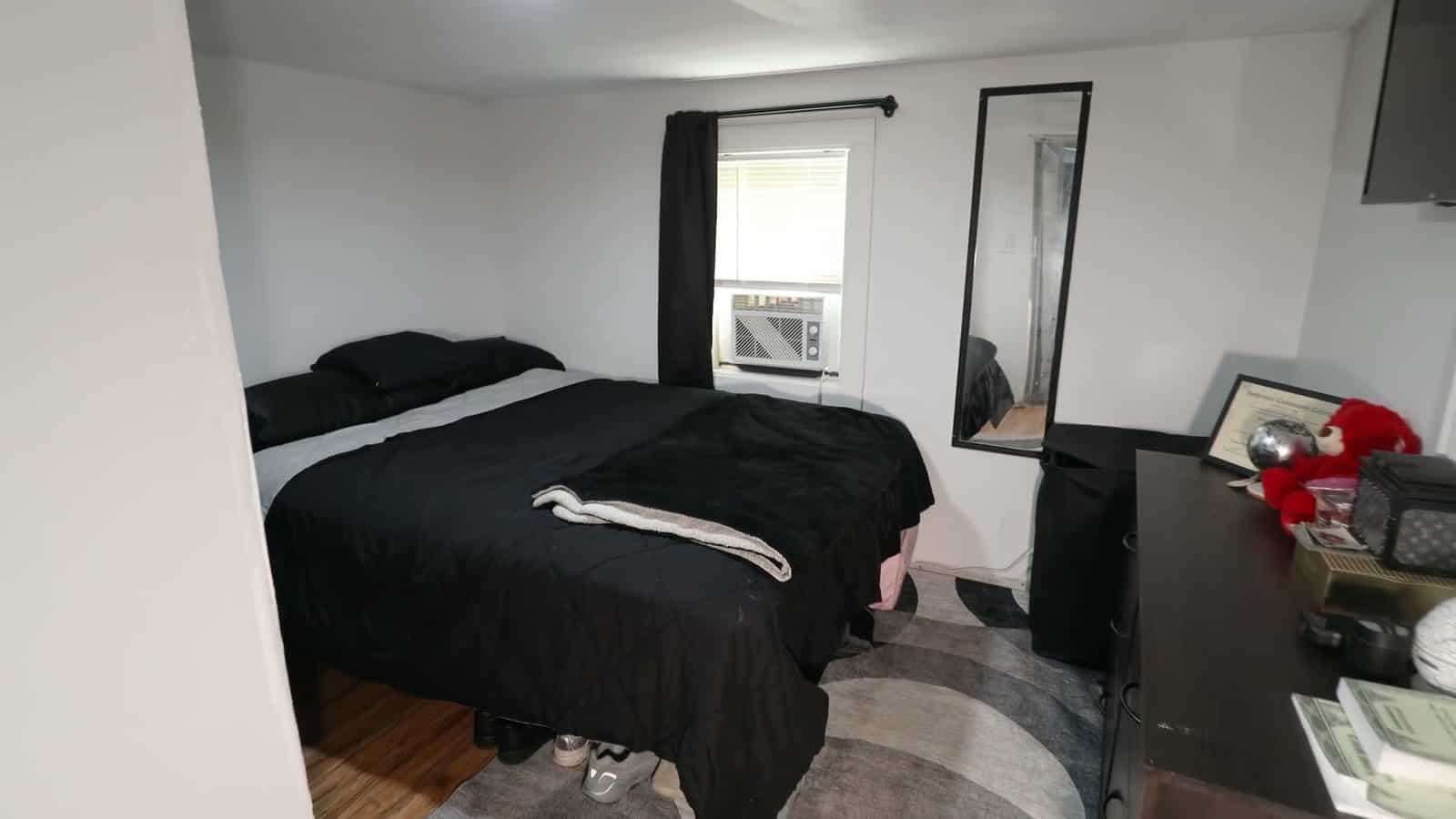 Bedroom in a shed conversion with a queen bed in black bedding, a window air conditioner behind black curtains, a full-length mirror and a wood dresser