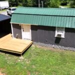 Dark-gray gambrel shed conversion with a green metal roof, white entry door, white lattice skirting, a window air conditioner, and a new wood deck running to a curtained gazebo