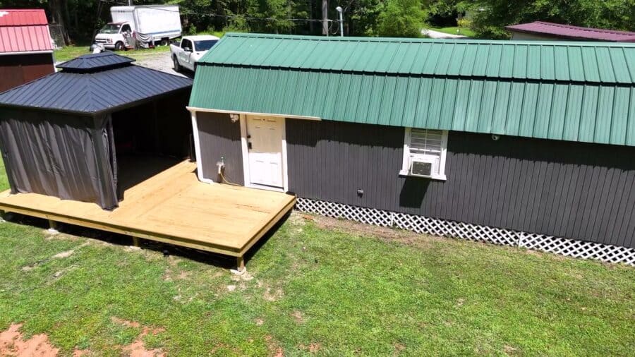 Dark-gray gambrel shed conversion with a green metal roof, white entry door, white lattice skirting, a window air conditioner, and a new wood deck running to a curtained gazebo