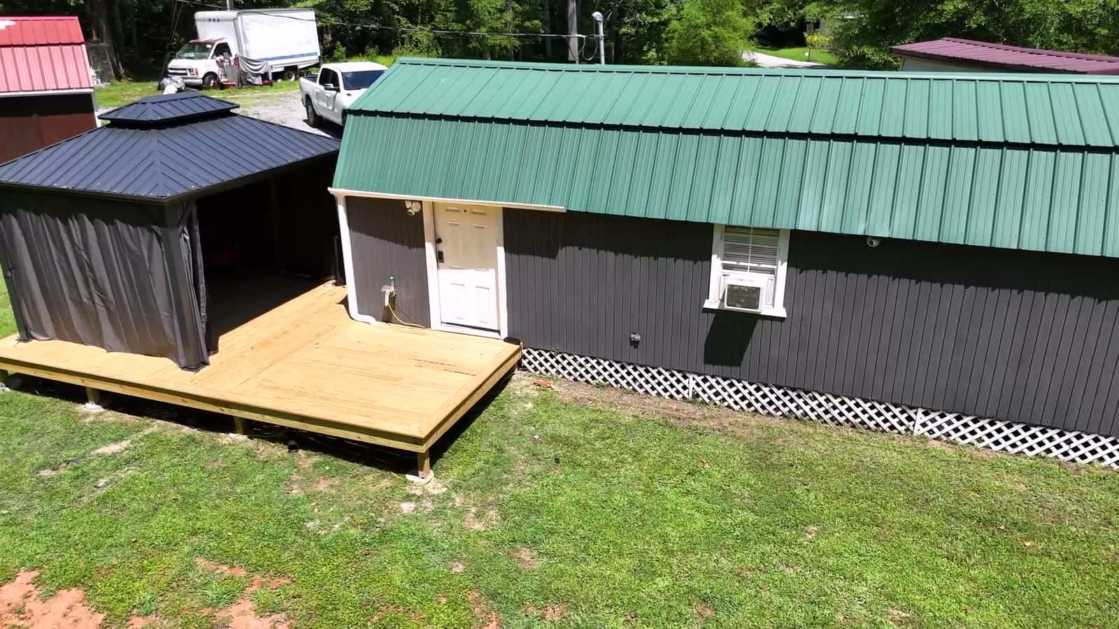 Dark-gray gambrel shed conversion with a green metal roof, white entry door, white lattice skirting, a window air conditioner, and a new wood deck running to a curtained gazebo
