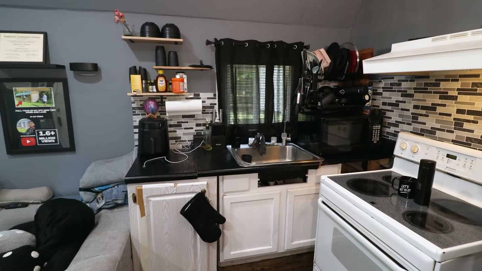 Kitchen in a shed conversion with a black counter, stainless sink, white electric range and hood, tile backsplash, floating wood shelves and a curtained window
