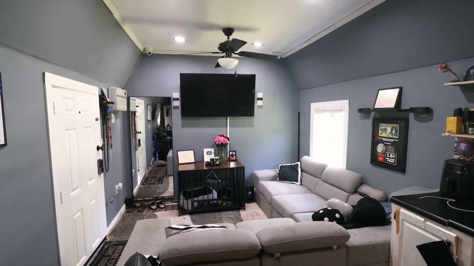 Living area of a shed conversion with gray walls, a wall-mounted TV, ceiling fan, gray sectional sofa, dog crate console and the kitchen counter at right