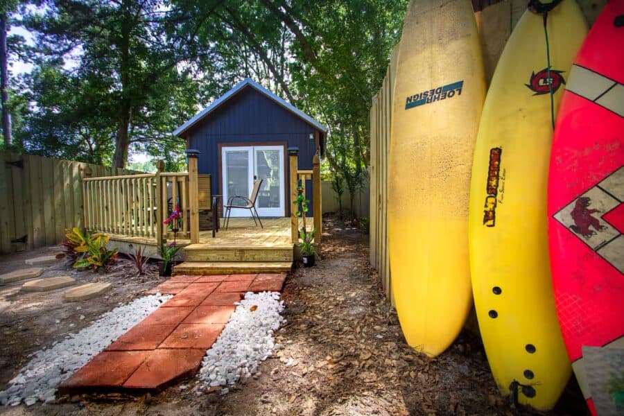 Cozy tiny house with a small deck, surrounded by trees, and surfboards leaning against a fence in a.