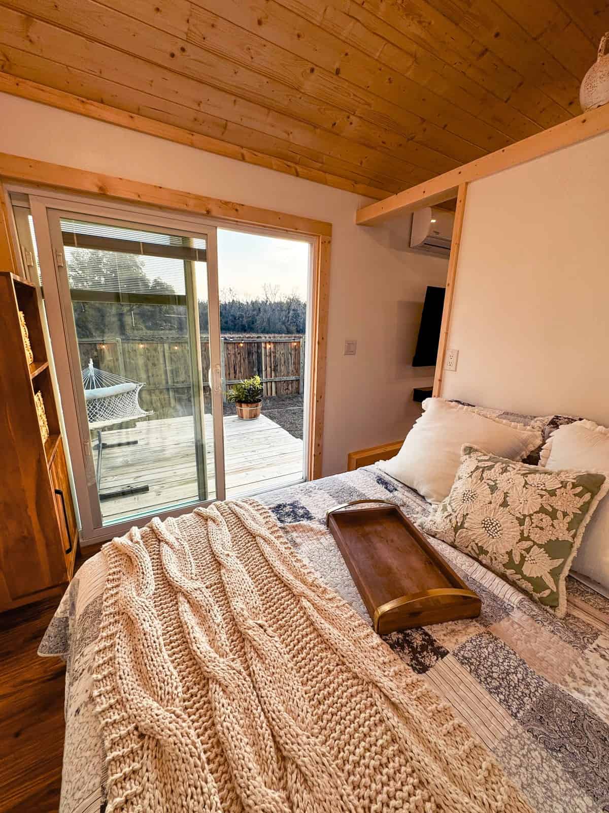 Main-floor queen bedroom in the Surya tiny house with sliding glass doors opening onto the deck, a mini-split, and a wall-mounted TV