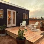 Long black Surya tiny house at dusk with a metal roof, French doors opening to a wood deck with Adirondack chairs, and an open field with a windmill behind