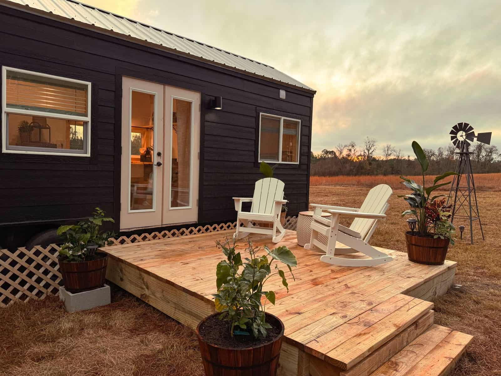 Long black Surya tiny house at dusk with a metal roof, French doors opening to a wood deck with Adirondack chairs, and an open field with a windmill behind