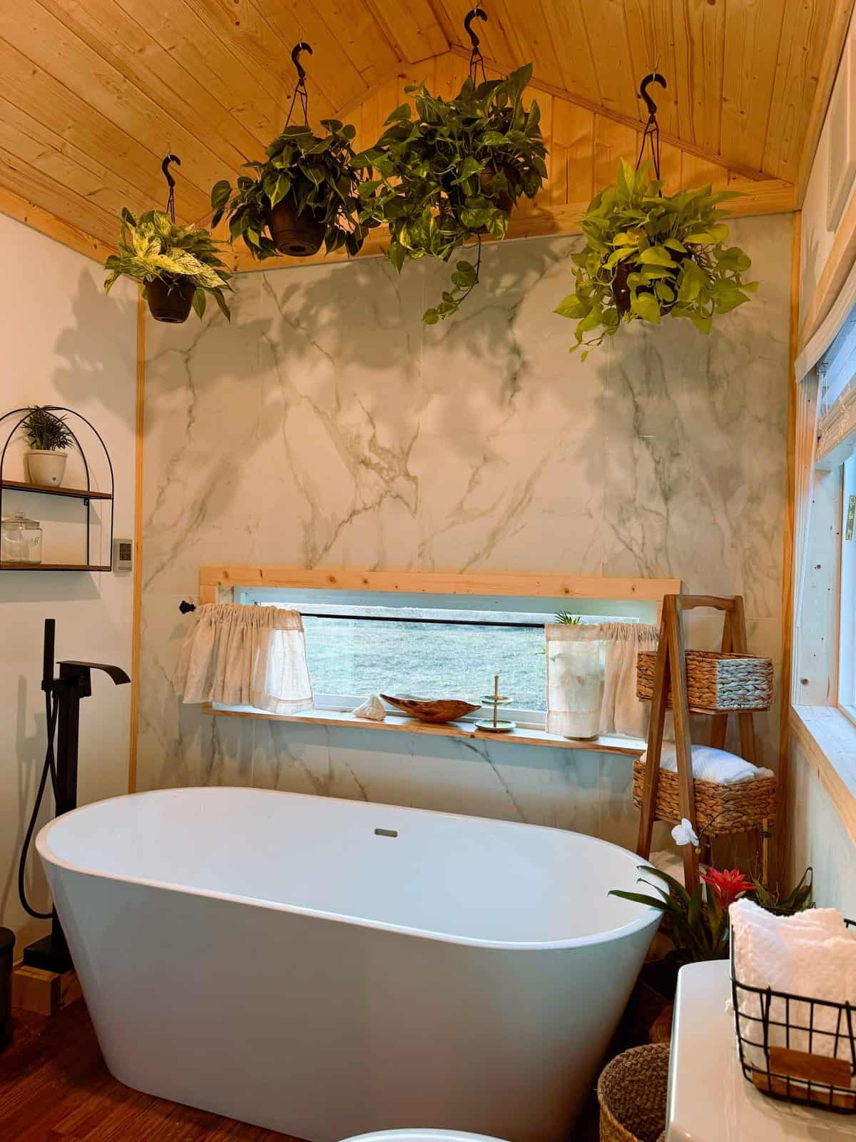 Spa-like bathroom in the Surya tiny house with a freestanding soaking tub, a marble-look wall, hanging plants, and a window