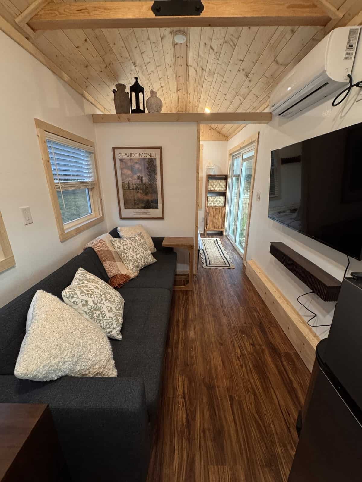Living room of the Surya tiny house with a sofa, a wall-mounted TV, a mini-split, and a vaulted pine ceiling, looking toward the bedroom