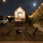 Tiny house with large glass front and outdoor seating area at night.
