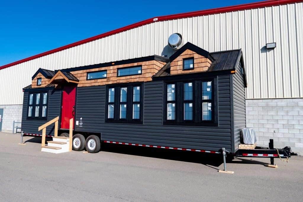 The Daphne park model by Teacup Tiny Homes with black lap siding, cedar shake dormers and a red front door