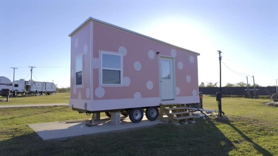 Small pink tiny house on wheels with polka dot pattern and front steps, set in open grassy area.