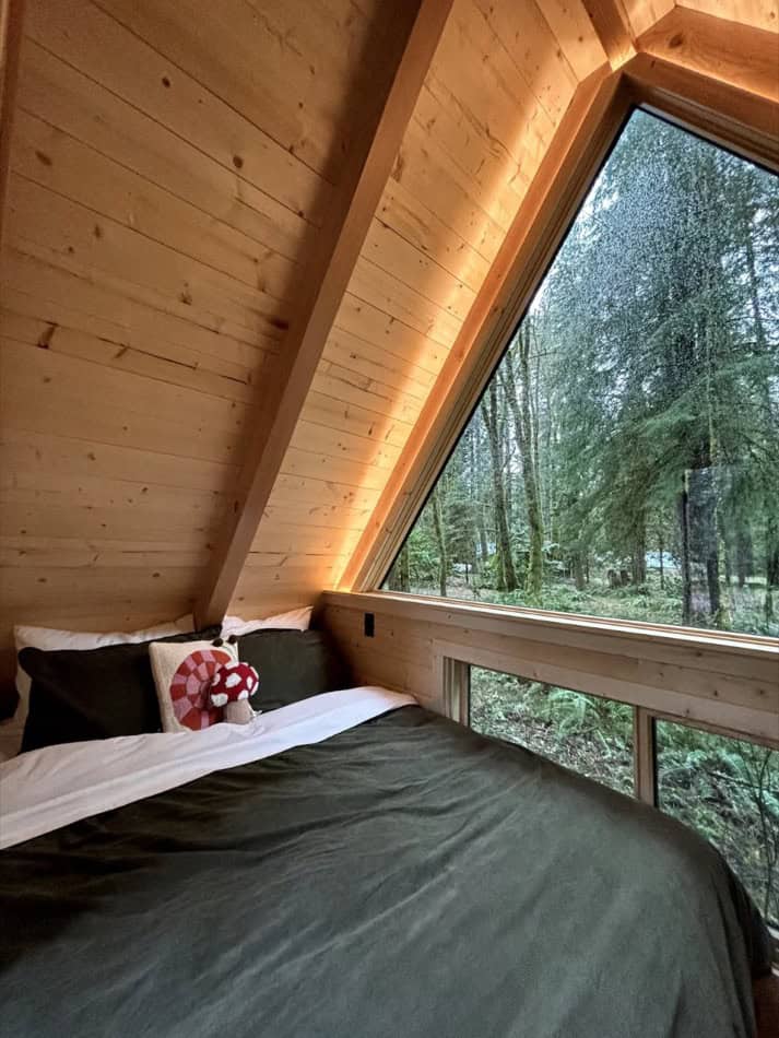 Interior of a tiny A-frame cabin with a cozy bed and large window overlooking a forest.
