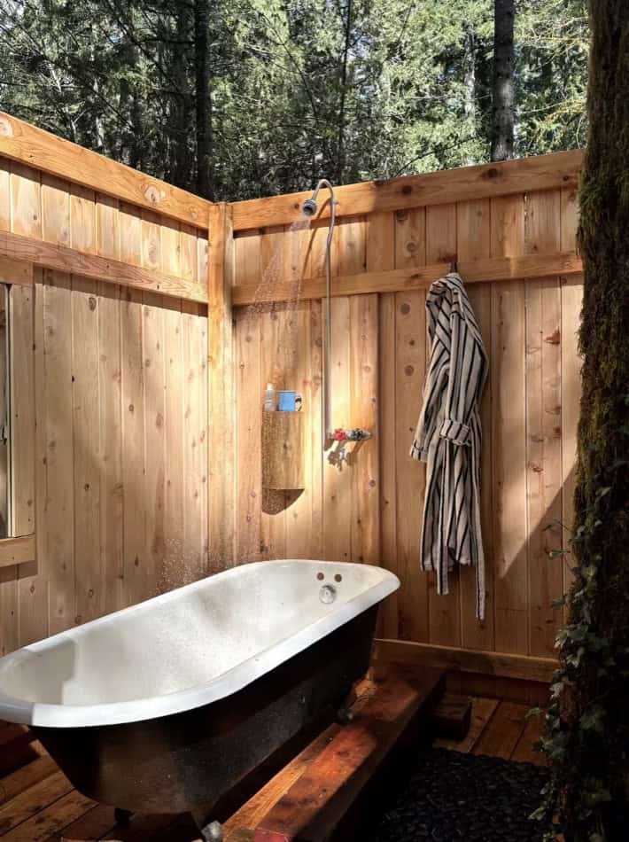 Outdoor bathtub with rustic wooden fencing and shower setup in a tiny cabin setting.