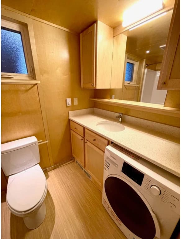 Efficient tiny house bathroom featuring a washer, wooden cabinets, and small window for natural ligh.