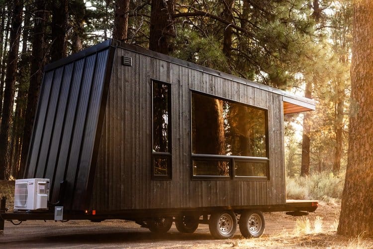 Modern black tiny house on wheels in a forest setting with large windows and natural sunlight.