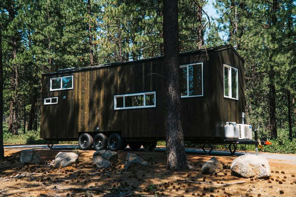 The Olympic tiny house by Spindrift Homes seen from its long side, with dark stained wood siding, white-trimmed windows, a mini-split unit, and a triple-axle trailer among pine trees
