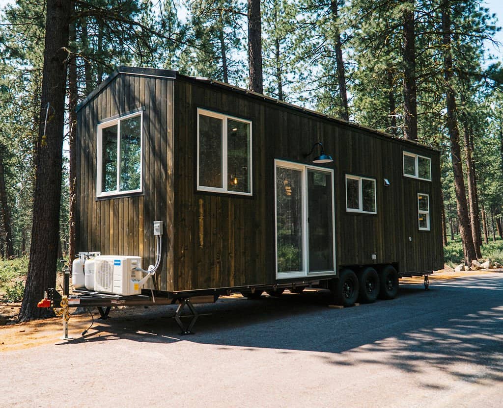 The Olympic tiny house by Spindrift Homes with dark wood siding and a metal roof on a triple-axle trailer in a pine forest