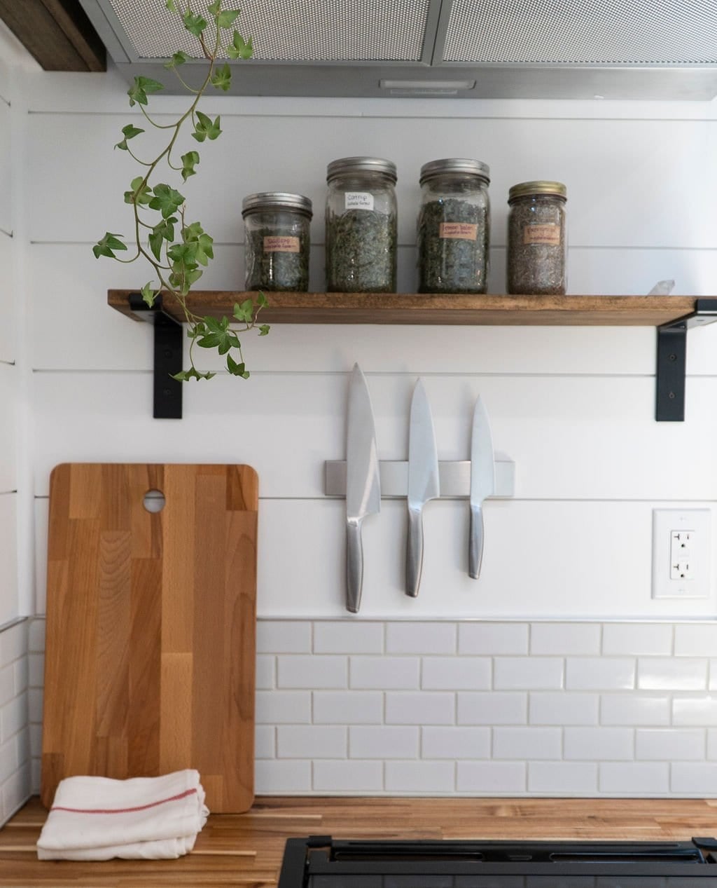 A reclaimed wood shelf with glass jars and a plant above a magnetic knife strip, subway tile, and acacia counter in the Olympic kitchen