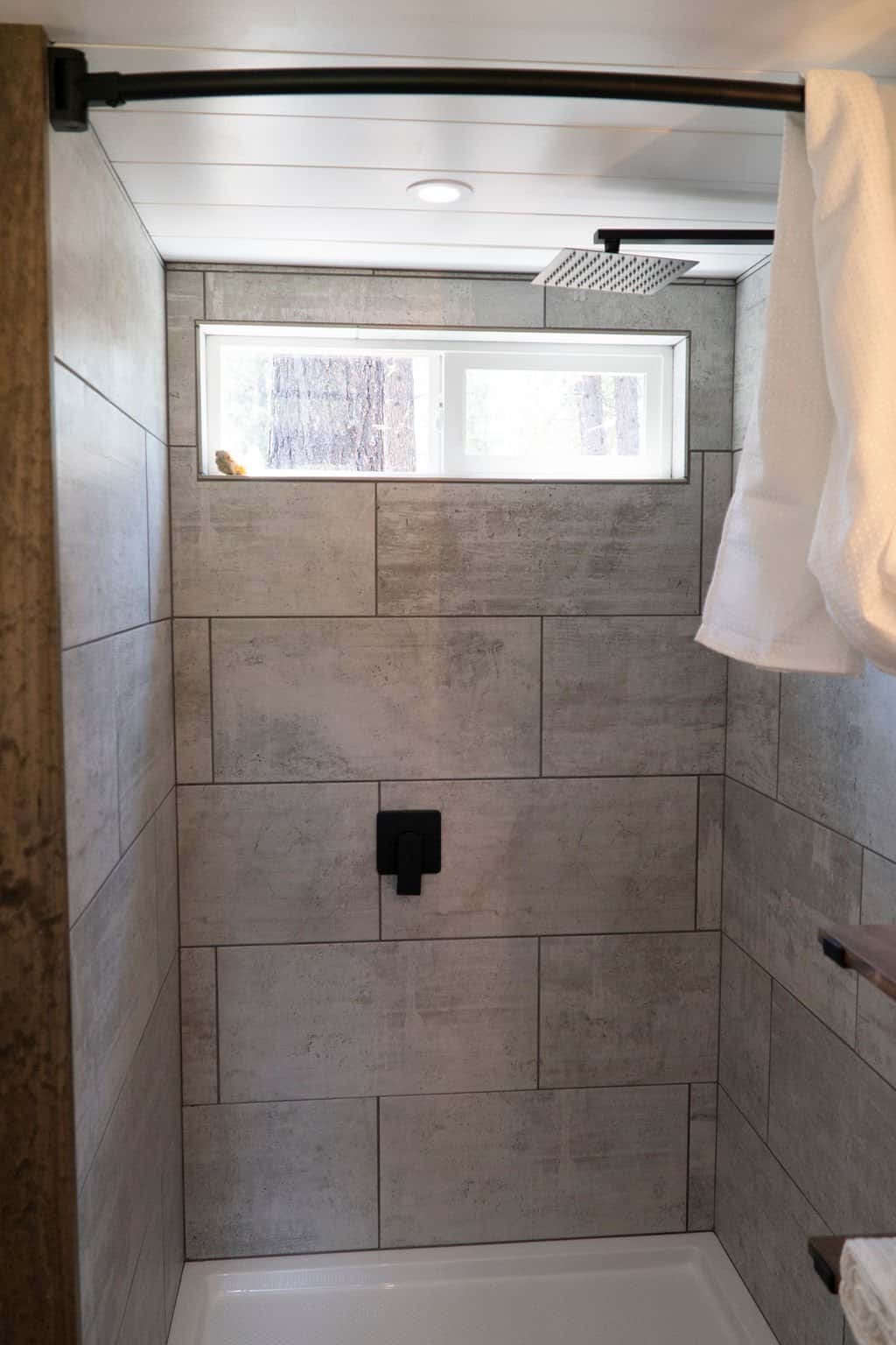 A fully tiled shower with concrete-look tile, a rainfall shower head, and a window in the Olympic tiny house