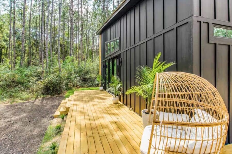 Modern tiny house with black exterior and wooden deck in a forest setting.