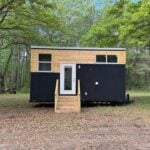 Tiny house on wheels with modern black and natural wood exterior, set in a lush green forest.