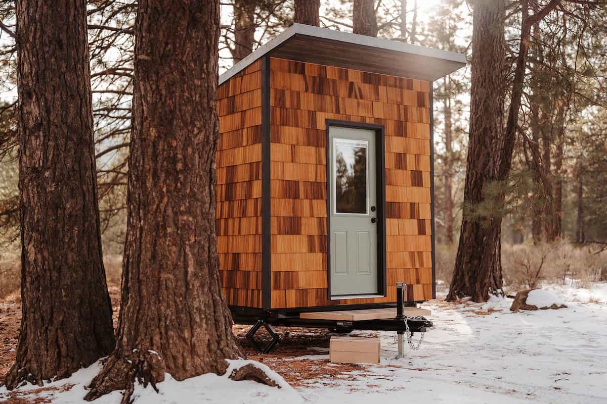 The River West mobile sauna by Spindrift Homes clad in cedar shingles with a slanted metal roof, parked among snowy pines
