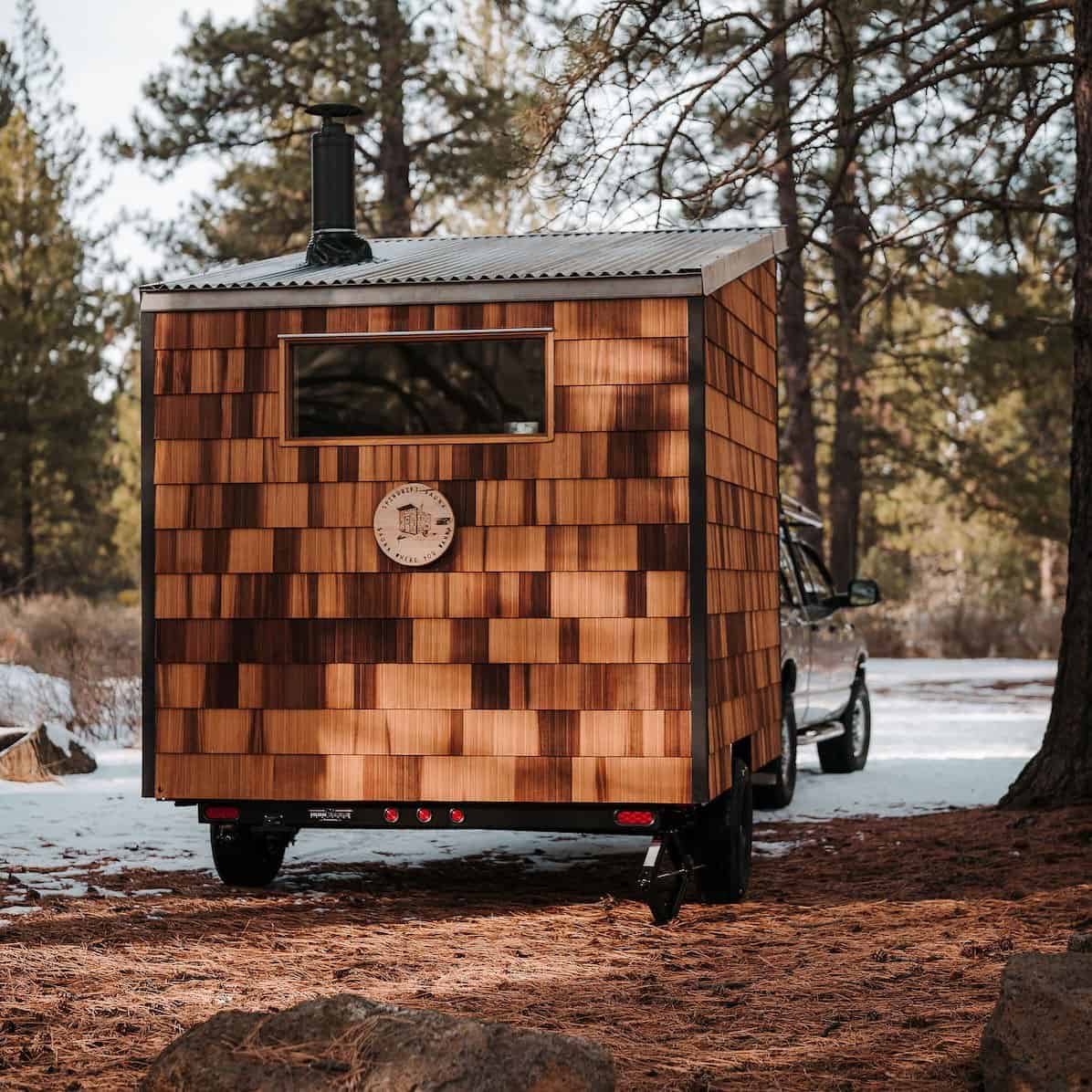 The River West cedar-shingle sauna hitched to a truck, showing its towable trailer and chimney