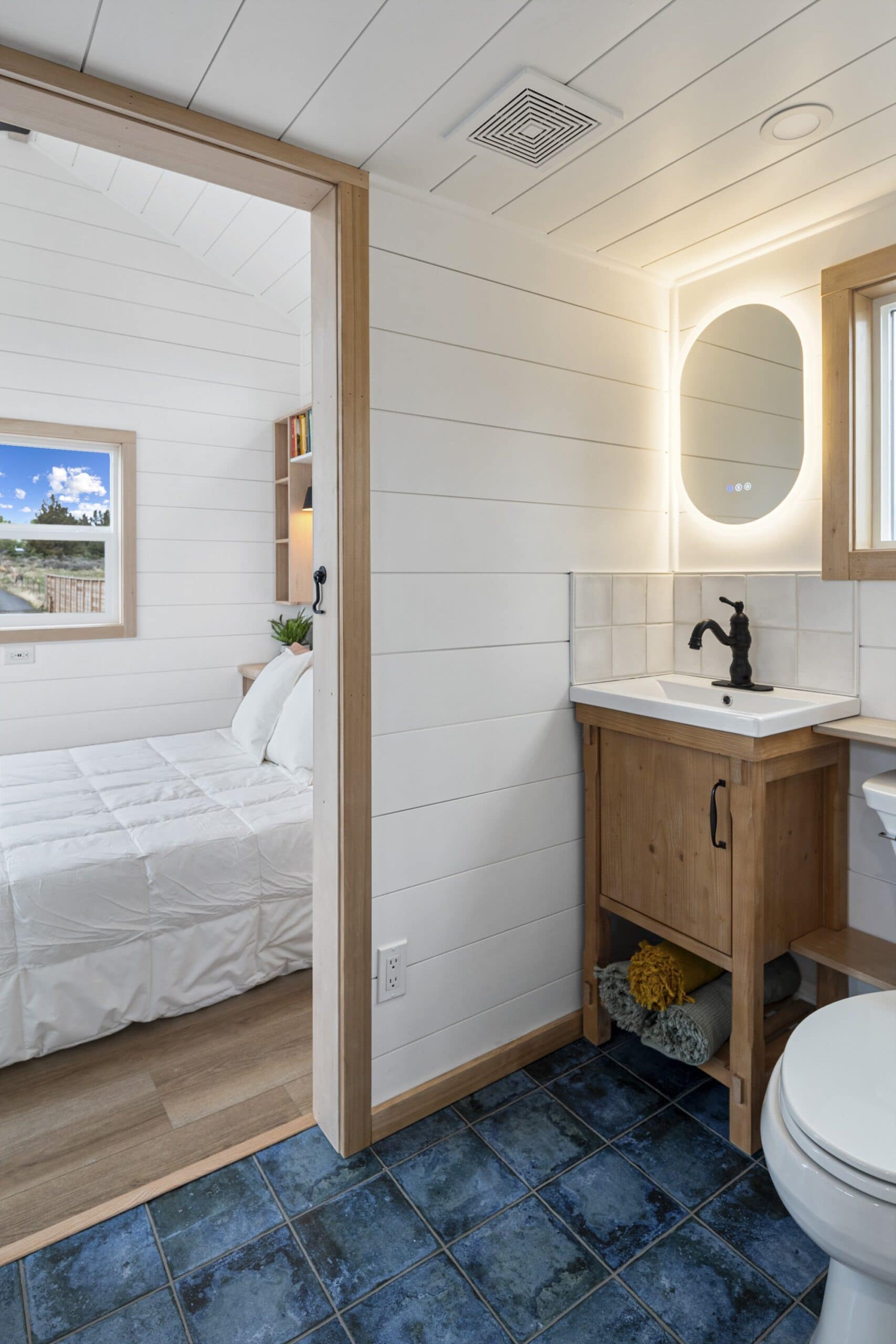 The Shasta bathroom with a wood vanity, vessel sink, illuminated mirror, and blue tile floor, with the bedroom beyond