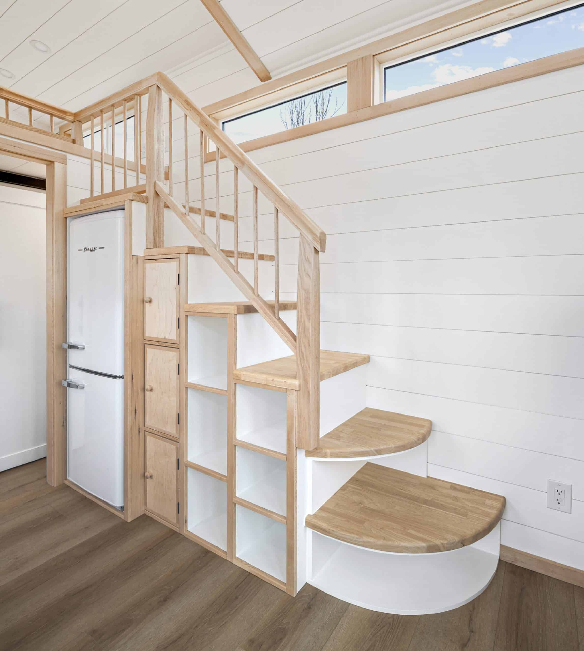 The Shasta interior showing the white Classic refrigerator, oak staircase, wood stove, daybed, and kitchen under a vaulted ceiling