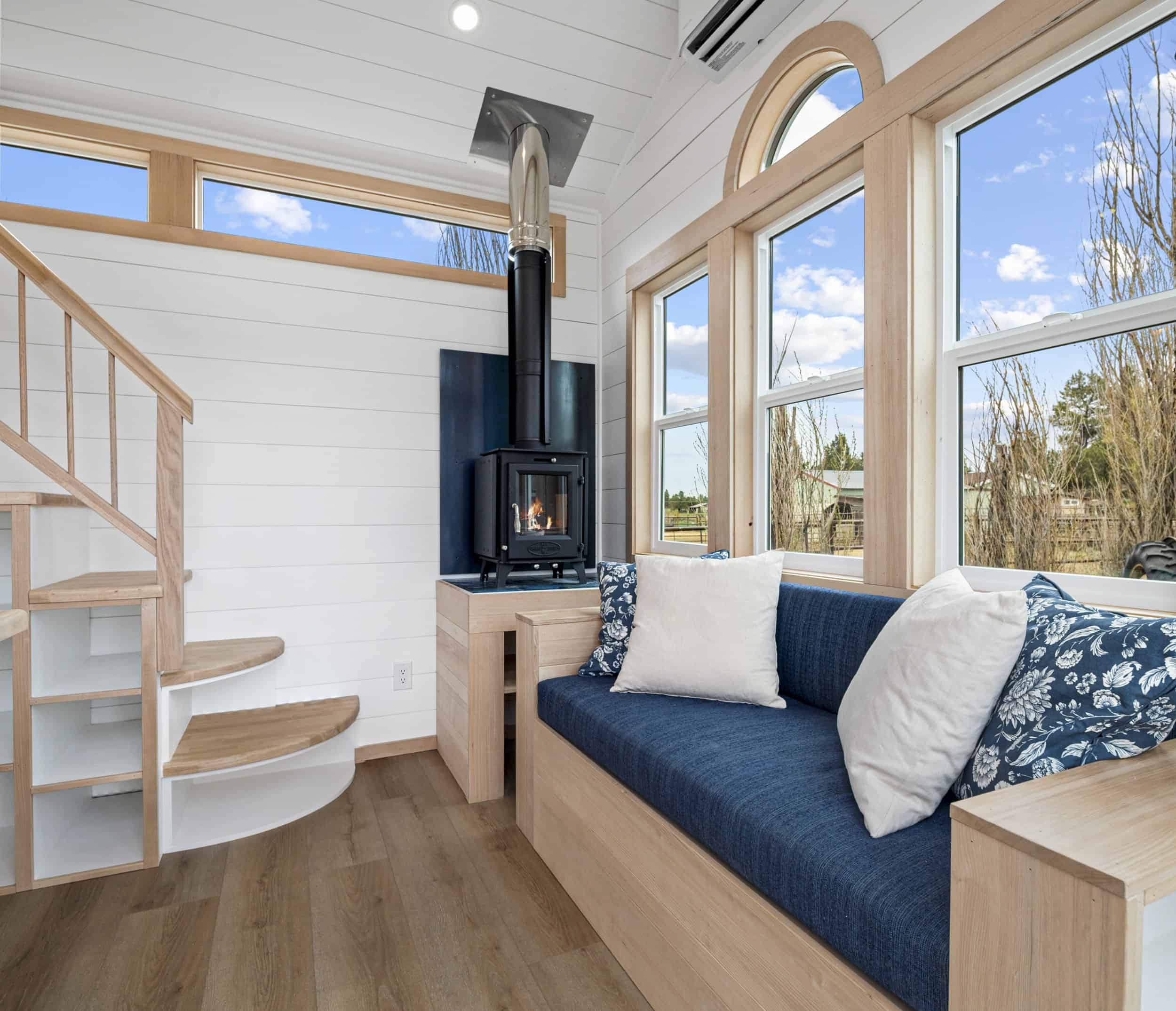 The Shasta living room with a wood-burning stove, a blue storage daybed, an arched window, and a curved oak staircase