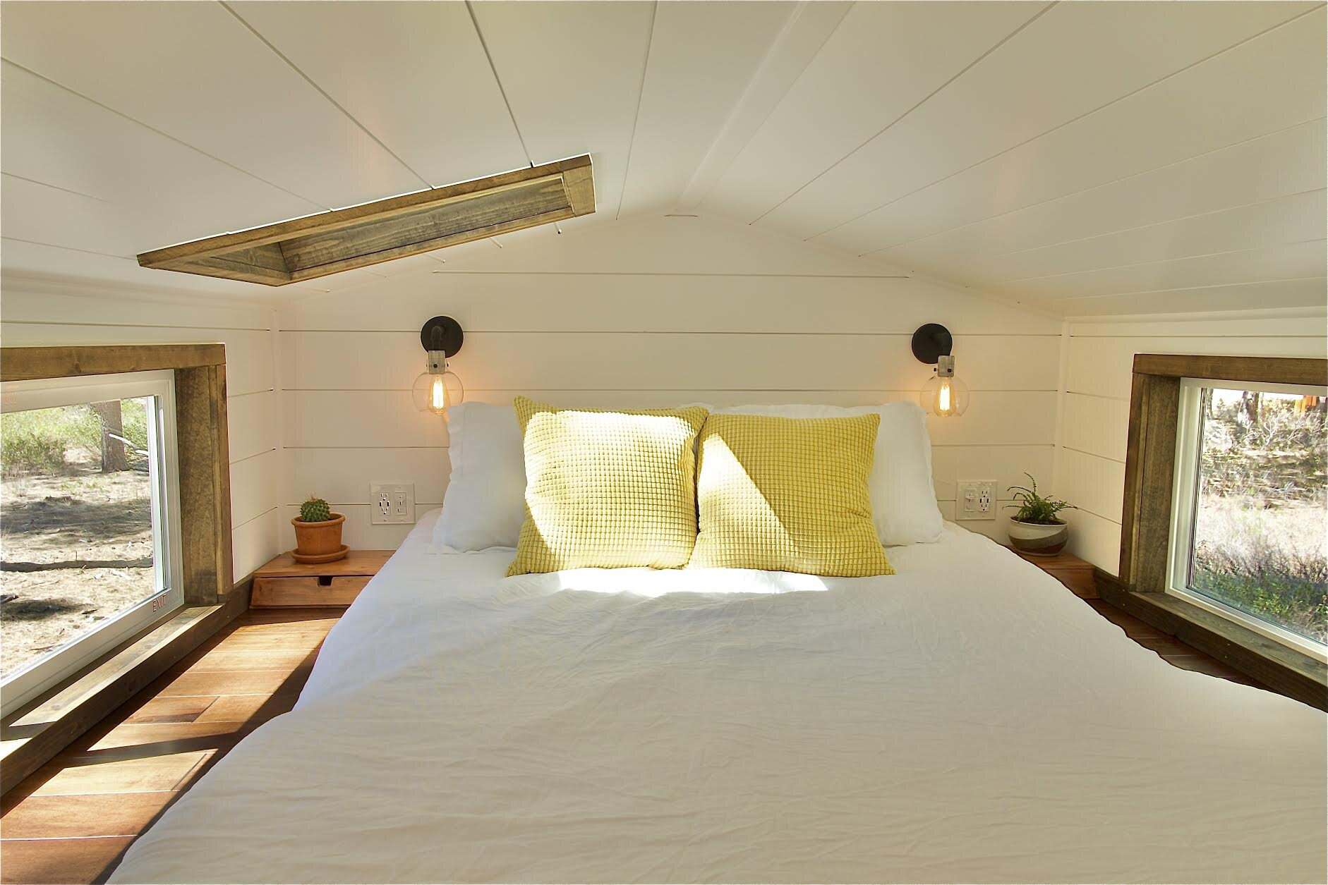 The Sonoma sleeping loft with a bed, yellow pillows, a skylight, and windows on both sides looking into the forest