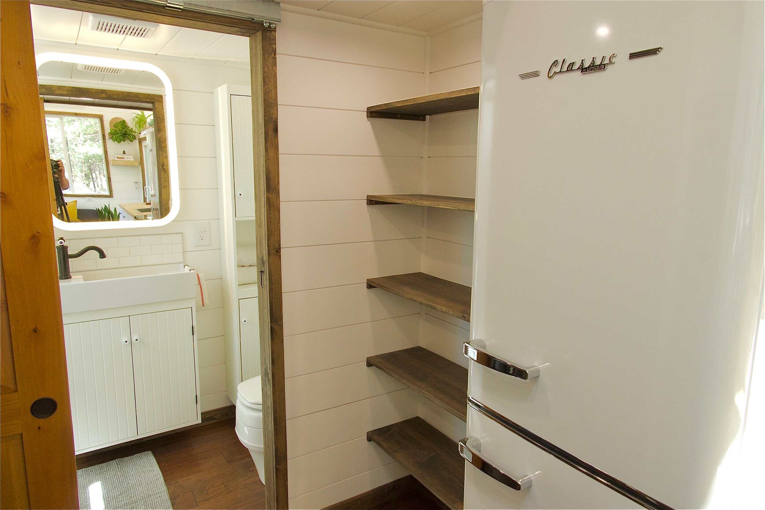 Open reclaimed-wood pantry shelving beside the refrigerator in the Sonoma tiny house, with the bathroom visible beyond