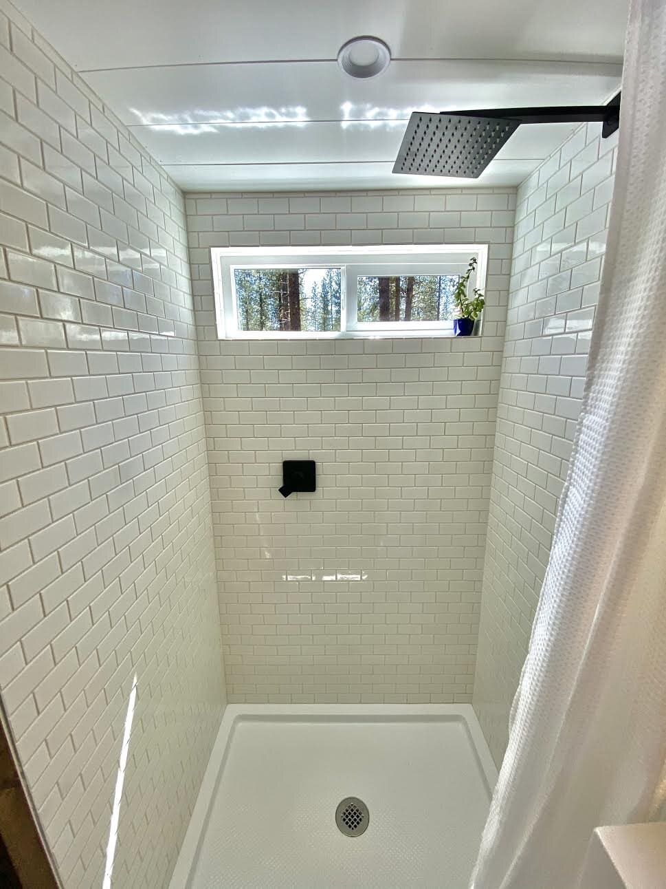A white subway-tiled shower with a rainfall shower head and a window in the Sonoma tiny house