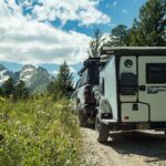 Off-grid tiny house with trailer on mountain trail surrounded by trees and mountains.