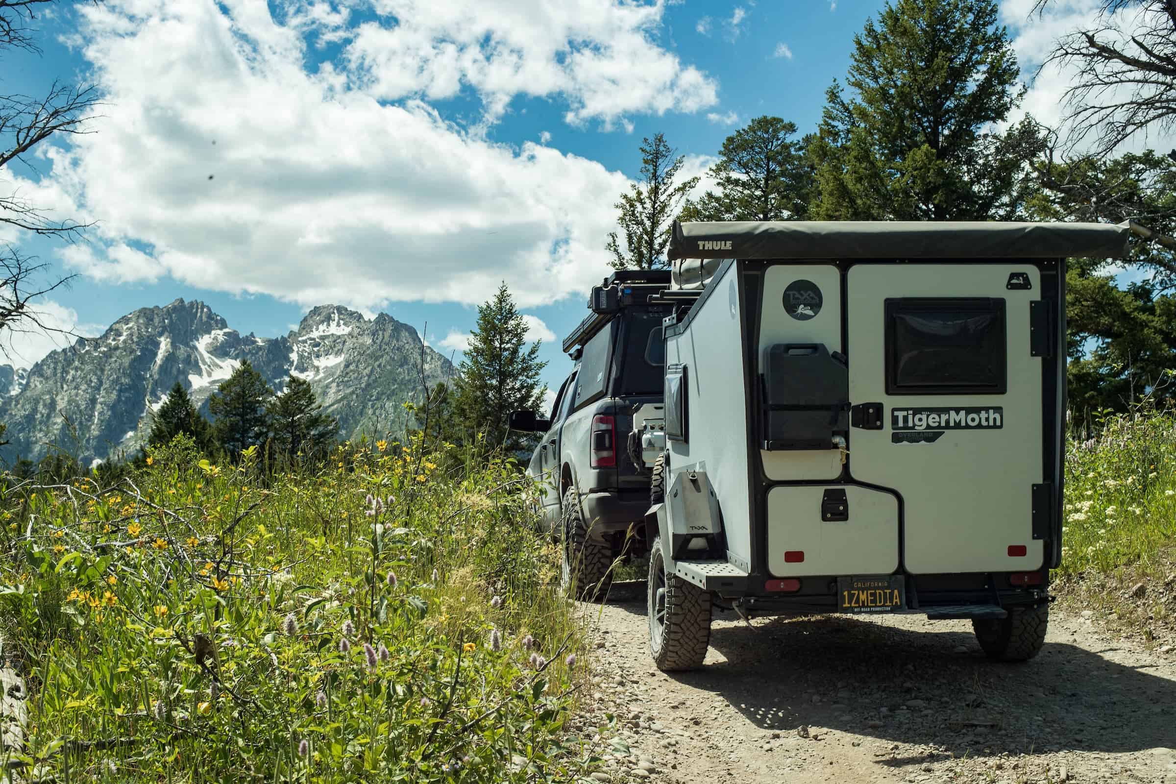 Off-grid tiny house with trailer on mountain trail surrounded by trees and mountains.