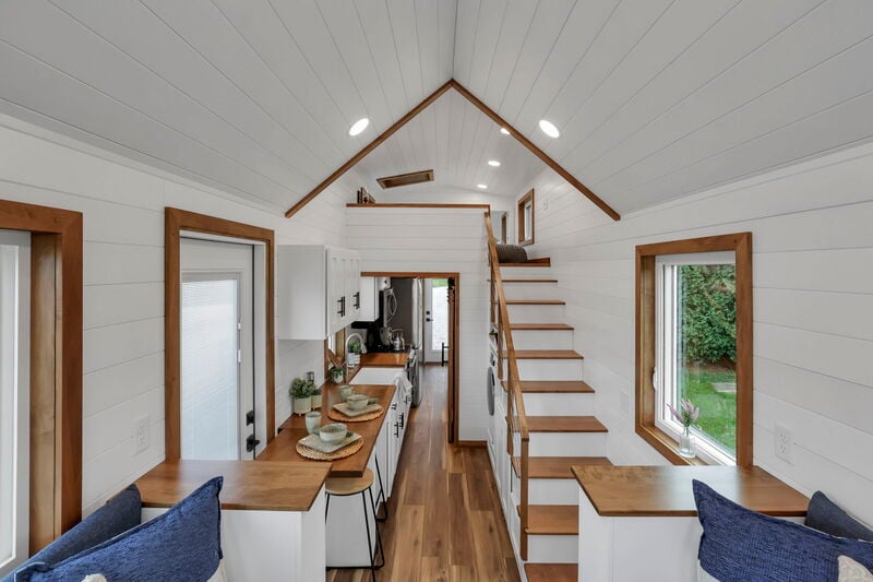 Tiny house interior with loft, white shiplap walls, wooden accents, and large windows.