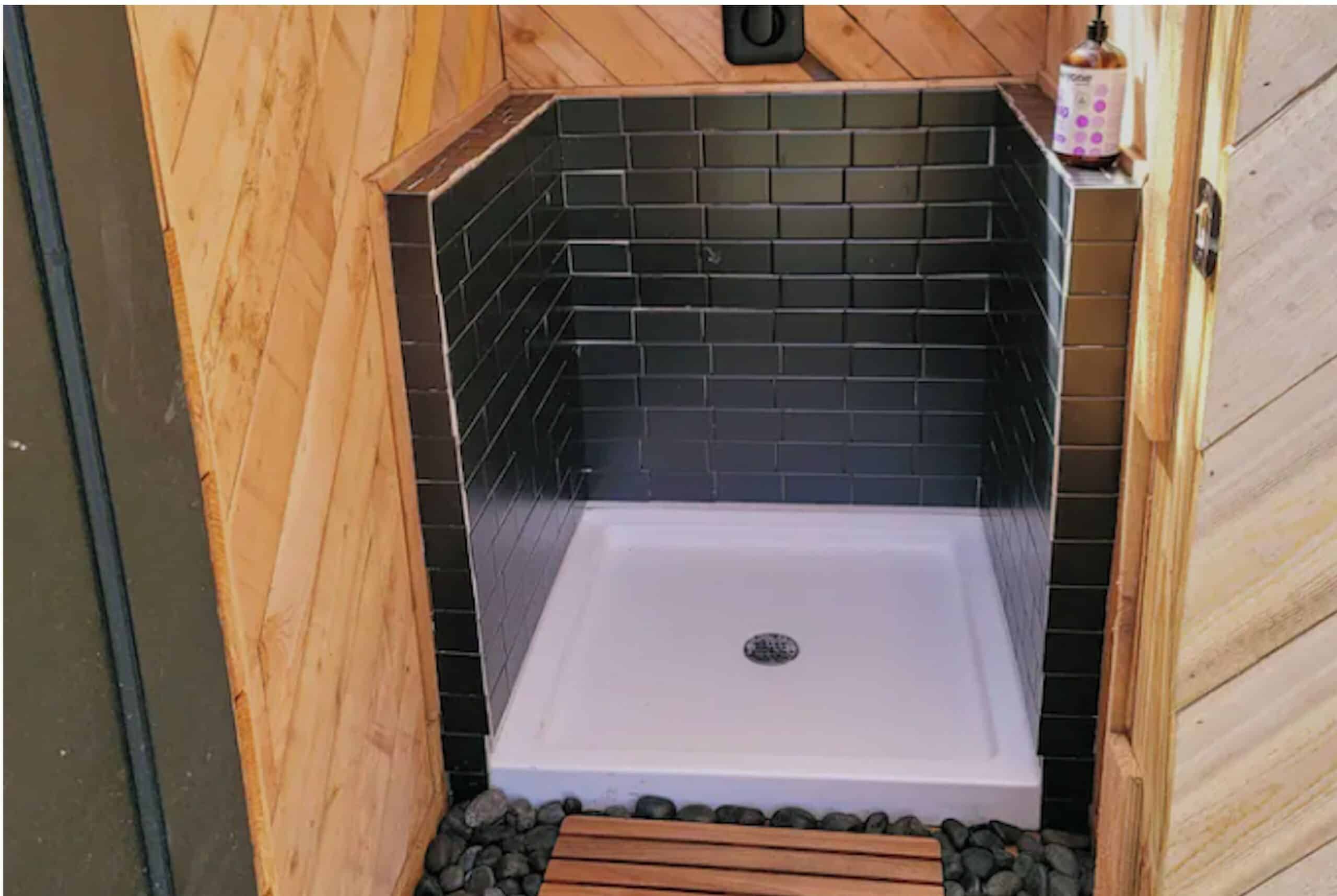 Black subway tile outdoor shower with river rock floor and teak mat at A-frame cabin near Austin