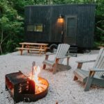 Tiny house cabin in a forest setting with outdoor fire pit and seating area.