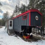 Compact tiny house in a wooded setting with snow-covered ground.