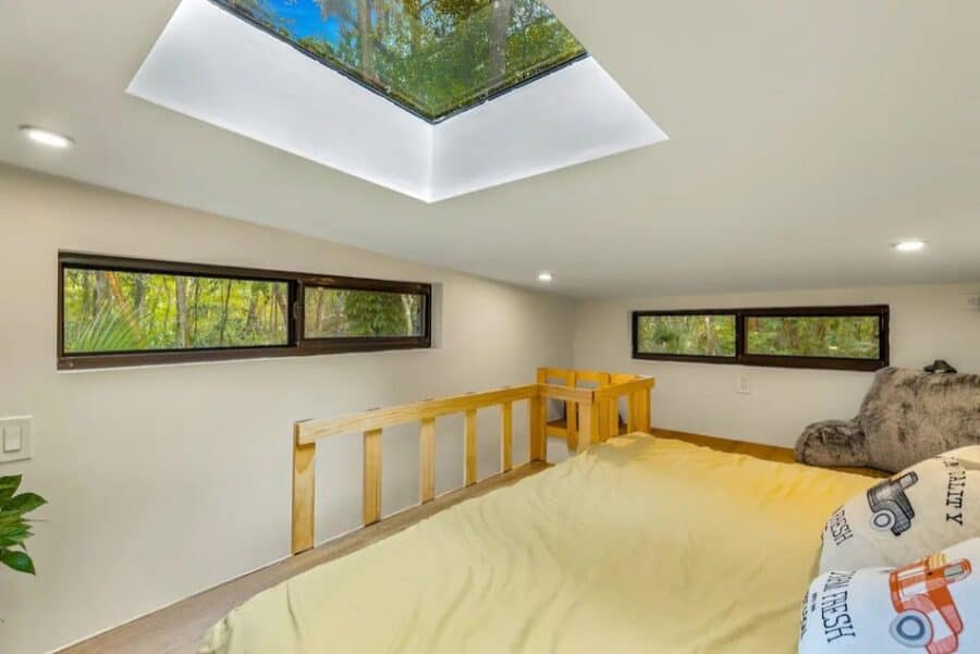 Bright tiny house bedroom with skylight and large windows overlooking lush greenery.