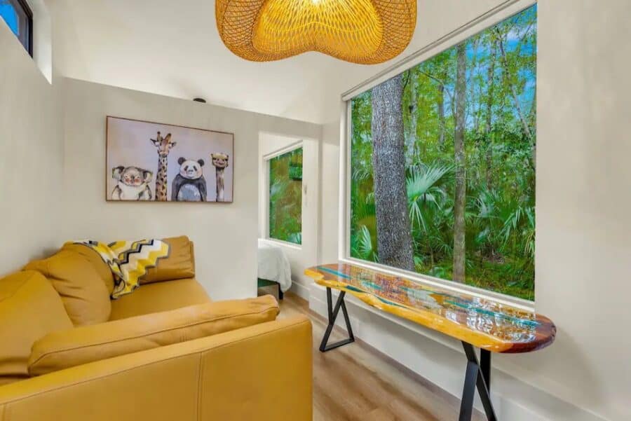 Bright interior of a tiny house with large windows showcasing lush forest scenery outside.