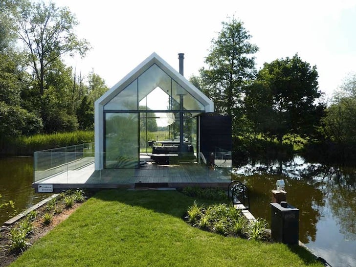Summer view of the glass cabin from the garden showing dark timber cladding and wooden deck by the lake