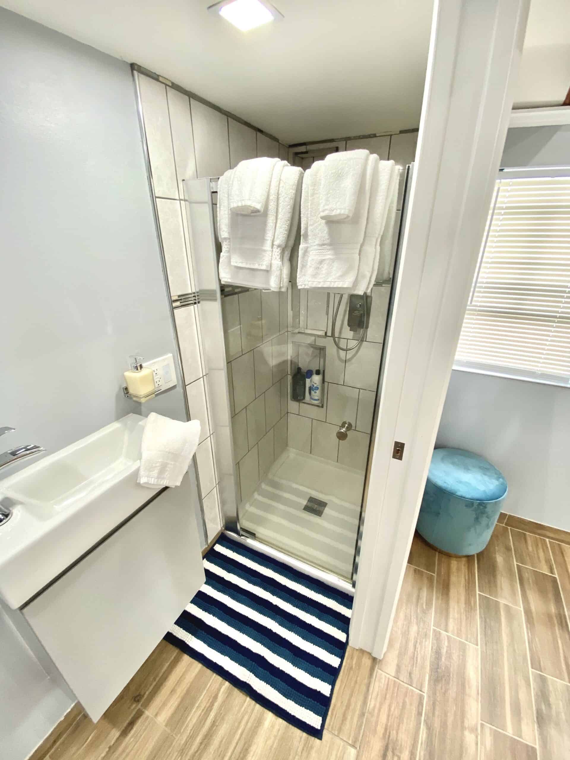 Tile shower with glass door, towels, and striped mat in the tiny cottage bathroom
