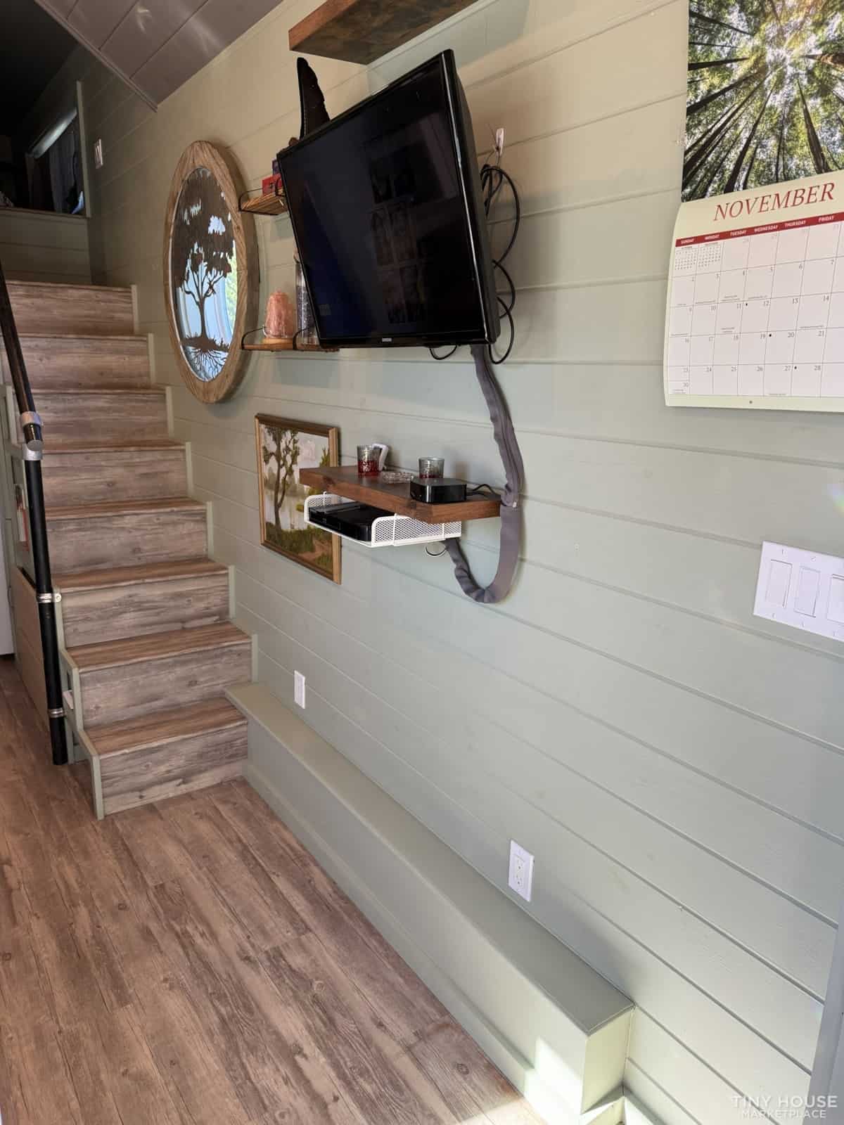 Living area with wall-mounted TV, tree artwork, staircase, and sage green shiplap walls