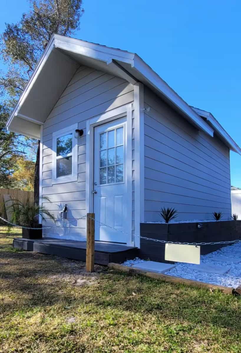 Small white tiny house with a gabled roof and front door, set in a sunny outdoor area.