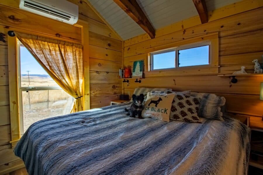 Cozy tiny house bedroom with wooden interior and large windows overlooking nature.