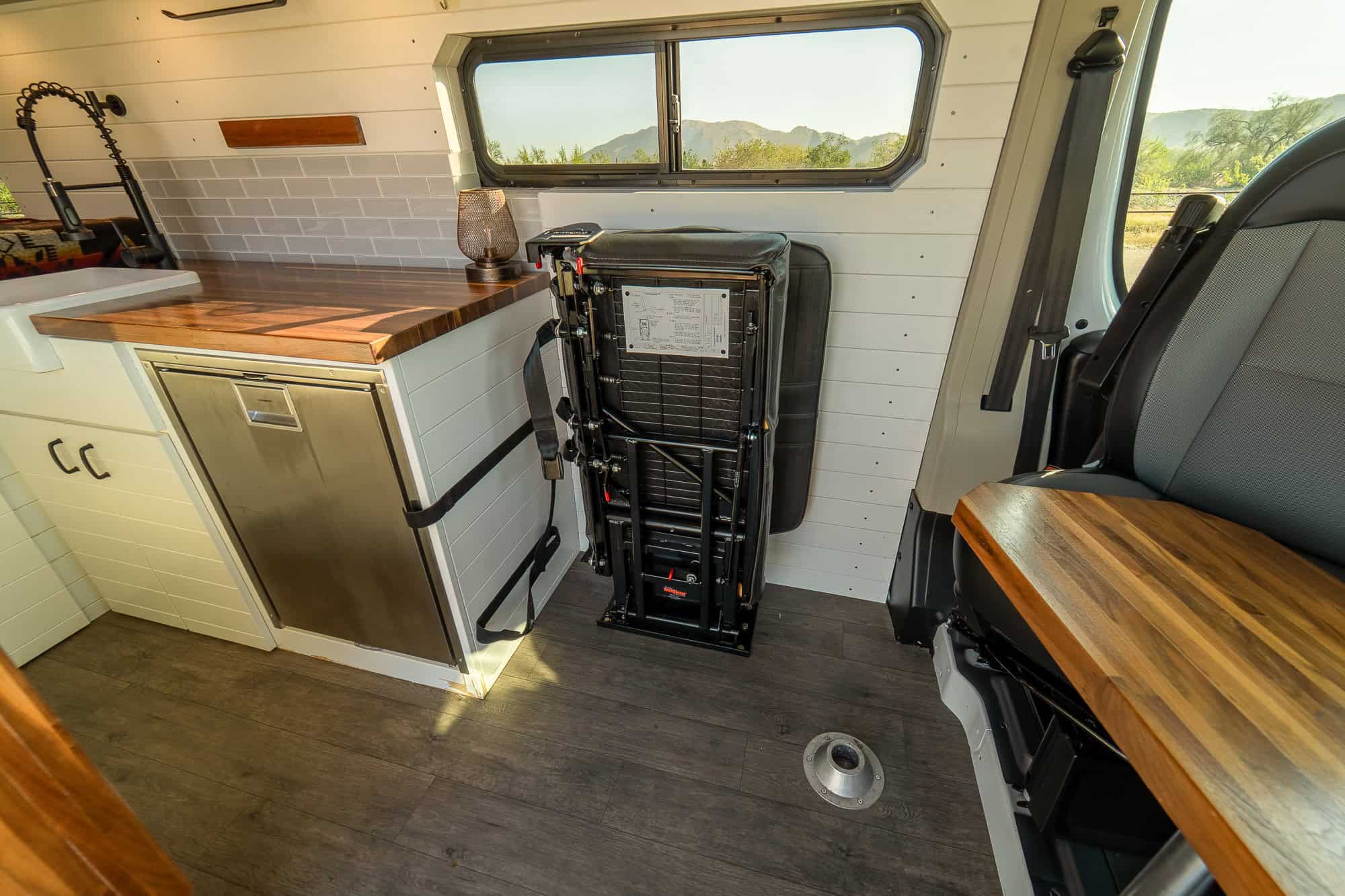 Folding jump seat stowed vertically against the wall of the ProMaster camper van next to a stainless Dometic fridge