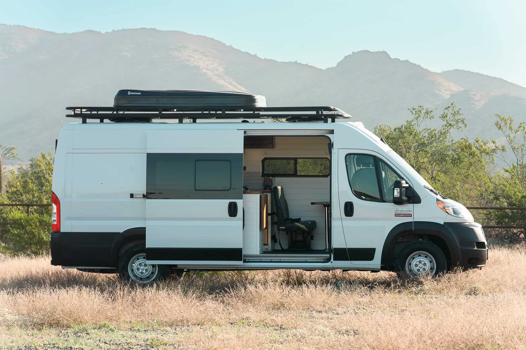 White 2023 Ram ProMaster camper van with black roof rack and rooftop tent parked in Arizona desert grass